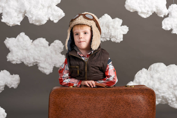 Als Flugzeugpilot verkleideter Junge steht mit altem Koffer zwischen den Wolken und spielt mit selbstgebasteltem Flugzeug - Foto, Bild