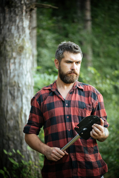 Brutal lumberjack with little spade wandering in wilderness. Ecologist with stylish beard exploring the woods. Young scientist working in nature, environment and ecology concept - Foto, Imagen