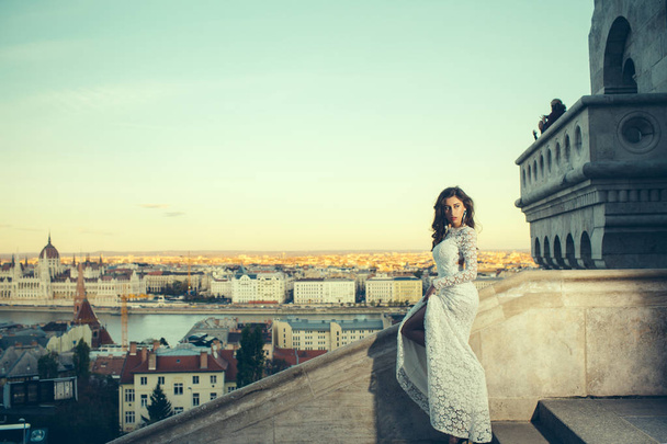 Girl with glamour look. Woman in white wedding dress on city view, fashion. Sensual woman with long hair on balcony, beauty. Bride with makeup. Fashion model style and hairstyle - Φωτογραφία, εικόνα