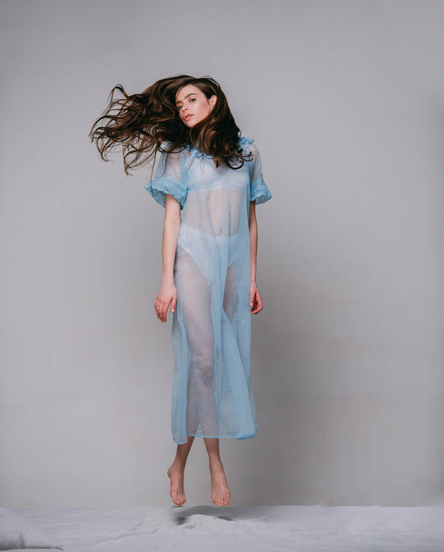 Lady in transparent blue nightie and white lingerie fly above bed, grey background. Woman with long curly hair wears tender pajama. Girl on calm face jump on bed. Femininity concept. - Photo, Image