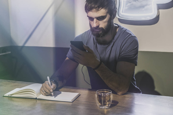 Young bearded man sitting in cafe and using mobile phone for listening digital music. Man making notes at note book. Horizontal. Blurred background. - Fotoğraf, Görsel
