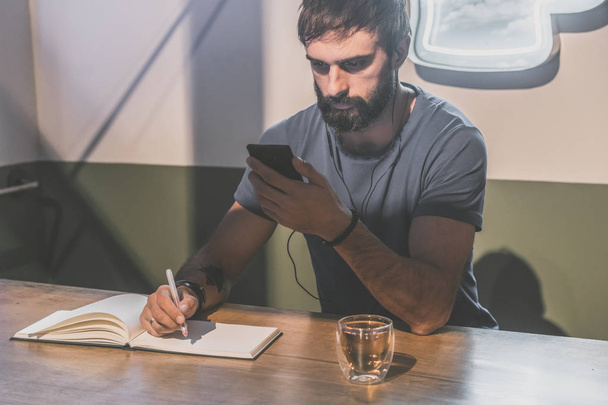 Young bearded man sitting in cafe with headphones. Men using mobile phone for listening digital music on smartphone. Man making notes at note book. Horizontal. Blurred background. Colors masks. - Foto, Bild