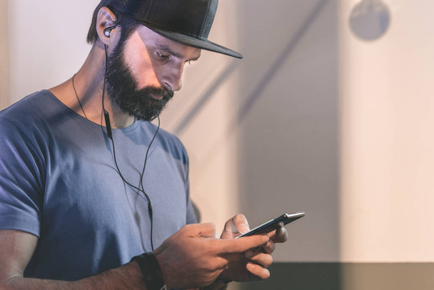 Bearded pensive man wearing black snapback cap standing on cafe with headphones. Men using mobile phone for listening digital music on smartphone.Blurred background. Horizontal. Visual effects. - Foto, Bild