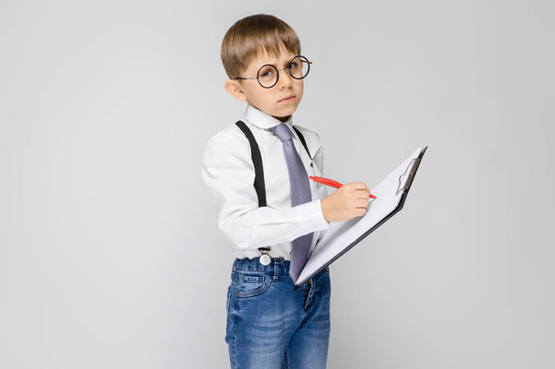 Um menino encantador com uma camisa branca, suspensórios, uma gravata e jeans leves fica em um fundo cinza. O menino segura uma caneta e lençóis para anotações
 - Foto, Imagem