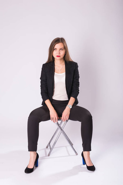 schöne junge Geschäftsfrau im schwarzen Anzug sitzt auf einem Stuhl auf weißem Hintergrund - Foto, Bild