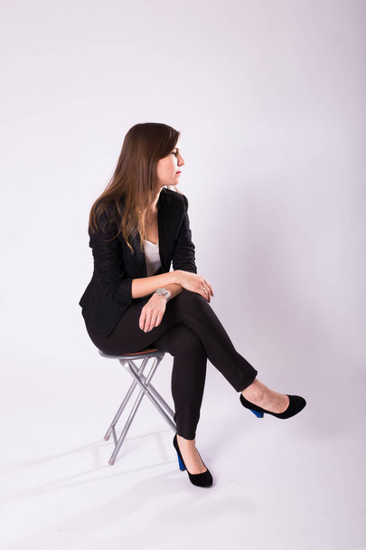 όμορφη νεαρή γυναίκα στο μαύρο κοστούμι κάθεται σε μια καρέκλα σε λευκό φόντο - Φωτογραφία, εικόνα