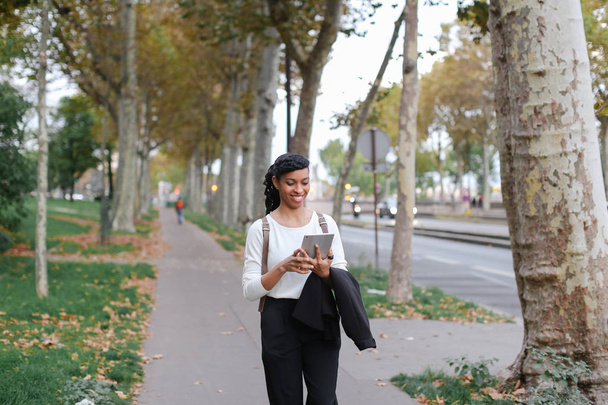 Bella ragazza nera che cammina dopo le lezioni con tablet sulla strada con foglie cadute
. - Foto, immagini