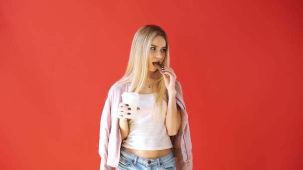 Girl on a red background, bites chocolate and drinks coffee - Séquence, vidéo