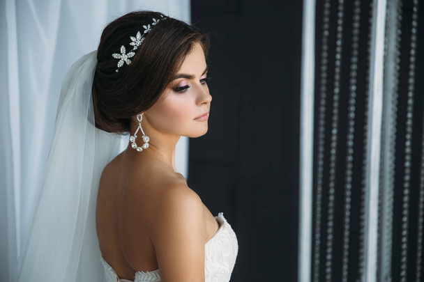 preparation of the bride on a wedding day. Beautiful brunette girl in a white luxury dress, with earrings, make-up and hairdo posing in a dark studio. Concept of marriage and family. - 写真・画像