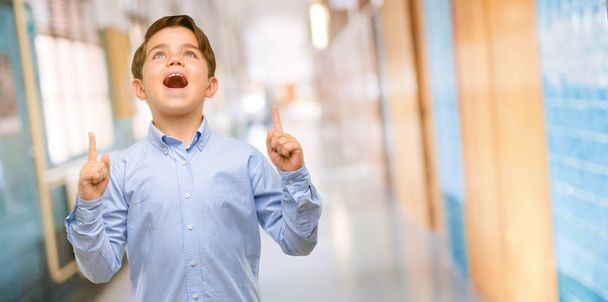 Όμορφος νήπιο παιδί με πράσινα μάτια, ευτυχισμένη και έκπληκτος επευφημίες εκφράζοντας wow χειρονομία που δείχνει προς τα επάνω στο διάδρομο του σχολείου - Φωτογραφία, εικόνα