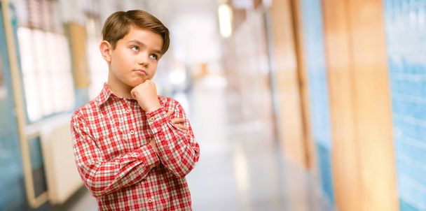Όμορφος νήπιο παιδί με πράσινα μάτια, σκέψης και κοιτώντας ψηλά εκφράζοντας αμφιβολίες και αναρωτιέμαι στο διάδρομο του σχολείου - Φωτογραφία, εικόνα