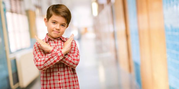 Όμορφος νήπιο παιδί με πράσινα μάτια ενοχλημένος με κακή στάση κάνοντας σήμα στοπ, διασχίζοντας τα χέρια, λέγοντας όχι, εκφράζοντας την ασφάλεια, άμυνα ή περιορισμό στο διάδρομο του σχολείου - Φωτογραφία, εικόνα