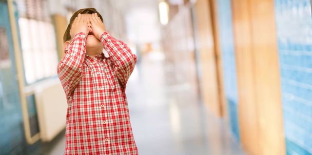 Όμορφος νήπιο παιδί με πράσινα μάτια αγχωτικό κρατώντας τα χέρια στο κεφάλι, κουρασμένοι και απογοητευμένοι στο διάδρομο του σχολείου - Φωτογραφία, εικόνα
