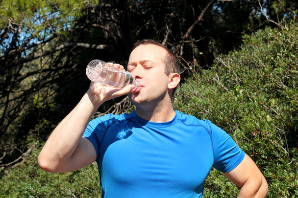 An athlete making a break during training, drinking fresh clean water from a bottle, rests and enjoys the clean air, with a beautiful landscape nature outdoors forest in background. Healthy lifestyle. - Photo, Image