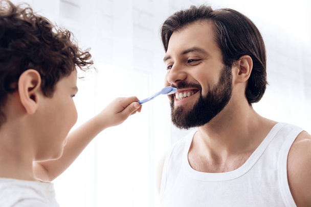 Little boy is brushing teeth of bearded man with toothbrush. - Foto, Imagen