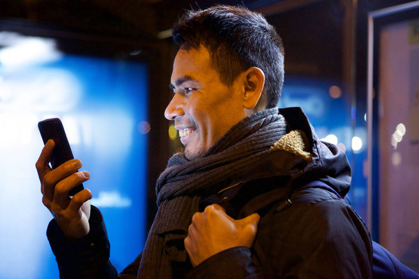 Close up side portrait of smiling mid adult asian man looking at his cell phone outdoors in night - Photo, Image
