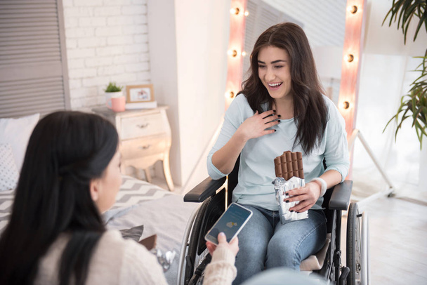 Lunch bite. Cheerful disabled woman and friend eating chocolate bar and communicating - 写真・画像