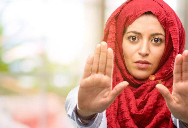 Mujer árabe joven usando hijab molesto con mala actitud haciendo señal de stop con la mano, diciendo que no, expresando seguridad, defensa o restricción, tal vez empujando
 - Foto, imagen
