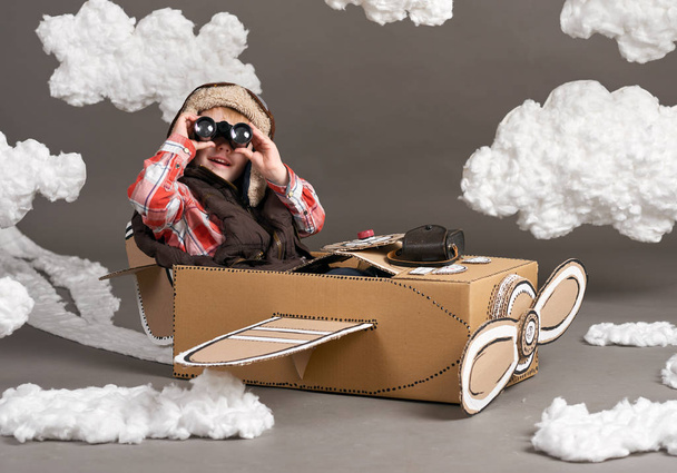 de jongen speelt in een vliegtuig gemaakt van kartonnen doos en droomt ervan om een piloot, wolken van katoen, wol op een grijze achtergrond - Foto, afbeelding
