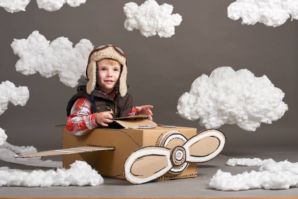 the boy plays in an airplane made of cardboard box and dreams of becoming a pilot, clouds of cotton wool on a gray background - Foto, Imagen