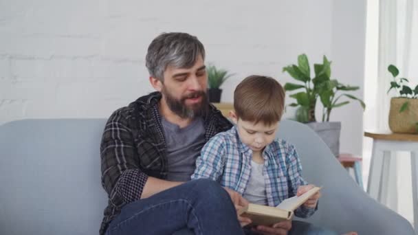 Caring father is teaching his little son curious preschooler to read. They are sitting together on sofa and reading book aloud wearing casual clothes. - Кадры, видео