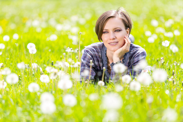 Beautiful woman enjoying dandelion field, lying down in the meadow of flowers and blowing seed away. Pretty girl relaxing outdoors on spring grass, having fun. Happy spring lady on green nature  - Photo, Image