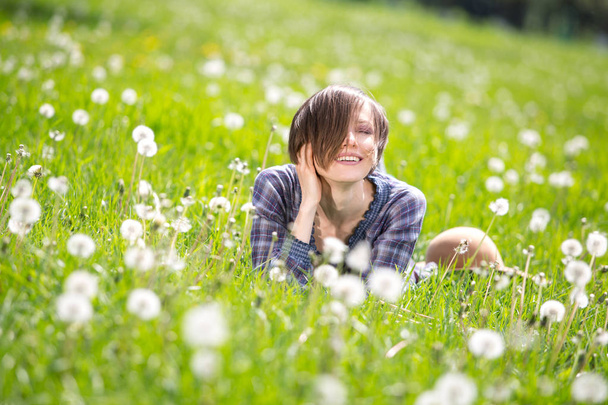Beautiful woman enjoying dandelion field, lying down in the meadow of flowers and blowing seed away. Pretty girl relaxing outdoors on spring grass, having fun. Happy spring lady on green nature  - Photo, Image