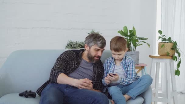 Adorable young boy and his caring father are using smartphone, talking and laughing together pointing at screen. Modern technologies and happy family concept. - Záběry, video