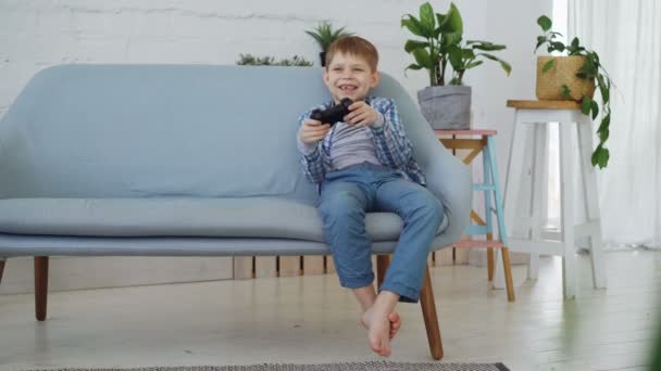 Video oyunu eğlenceli olan tek başına oynayan neşeli küçük çocuk evde kanepede oturuyor. Modern teknolojiler, mutlu çocukluk ve boş zaman zaman kavramı. - Video, Çekim