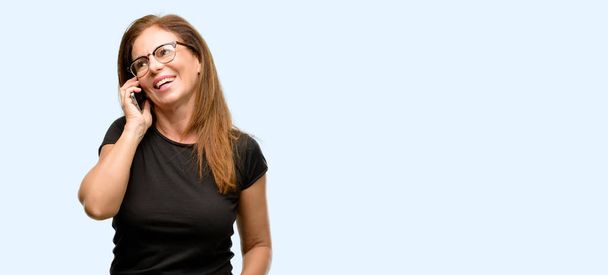 Donna di mezza età indossa camicia nera e occhiali felice parlando utilizzando uno smartphone cellulare isolato sfondo blu
 - Foto, immagini