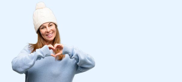 Женщина средних лет в шерстяной зимней шапочке счастлива, показывая любовь с руками в форме сердца, выражая здоровый и брак символ изолированный синий фон
 - Фото, изображение