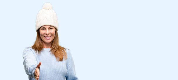 Middle age woman wearing wool winter cap holds hands welcoming in handshake pose, expressing trust and success concept, greeting isolated blue background - Фото, изображение