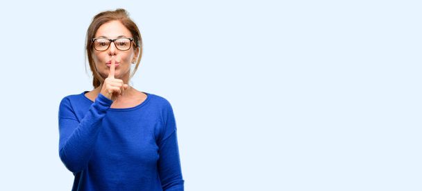 Middle age woman wearing wool sweater and glasses with index finger on lips, ask to be quiet. Silence and secret concept isolated blue background - 写真・画像