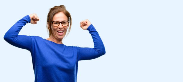 中年の女性はウールのセーターとメガネ幸せと興奮祝う勝利大成功、力、エネルギーおよび肯定的な感情を表現することを身に着けています。新しいジョブうれしそうな孤立した青い背景を祝う - 写真・画像