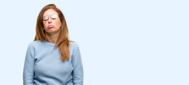 Женщина средних лет в шерстяном свитере и крутых очках со сонливым выражением лица, переутомленная и уставшая на синем фоне
 - Фото, изображение