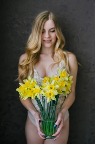  Hermosa mujer rubia sexy en lencería beige sexy con flores de primavera ramo de narcisos. Fokus selectivo en flores
 - Foto, Imagen