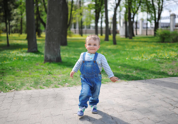 Cute baby boy with sweet smile, stands in a funny pose in green city park nature. - Photo, Image