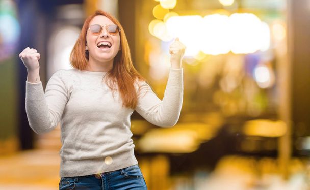 Красивая молодая рыжая женщина счастлива и взволнована, выражая победный жест. Успешная и торжествующая победа, торжествующая ночью
 - Фото, изображение