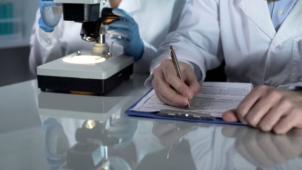 Travailleur de laboratoire remplissant des formulaires papier, son assistant regardant des échantillons au microscope
 - Photo, image