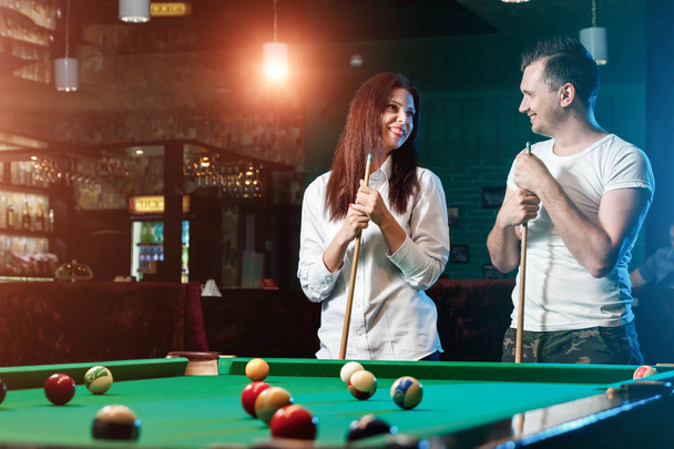A guy and a beautiful girl are playing billiards, a guy is teaching a girl to play billiards. Dark background. Pleasant pastime, family rest, entertainment, holiday. - Fotoğraf, Görsel