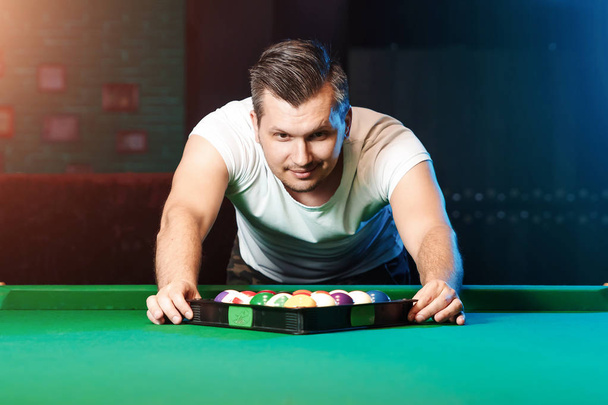 A man in a white T-shirt is playing billiards, a dark background. Pleasant pastime, entertainment, leisure, family holidays, games. - Фото, изображение