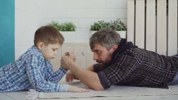 Cheerful father is teaching his little son arm wrestling, showing him hand position and pretenting to lose, boy is interested and concentrated on new activity. - Záběry, video
