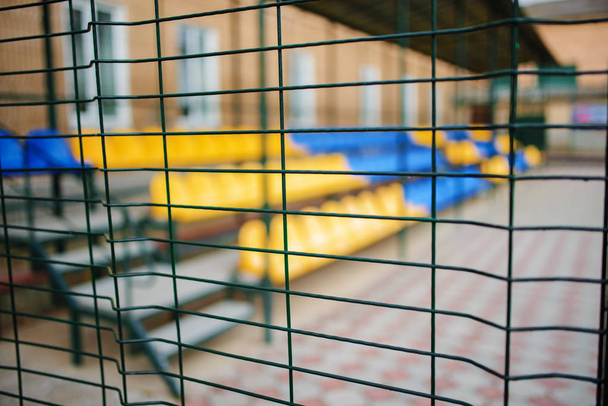 Feche a cerca metálica em forma de rede do arame em um fundo de assentos esportivos azuis e amarelos vazios do grande estande
 - Foto, Imagem