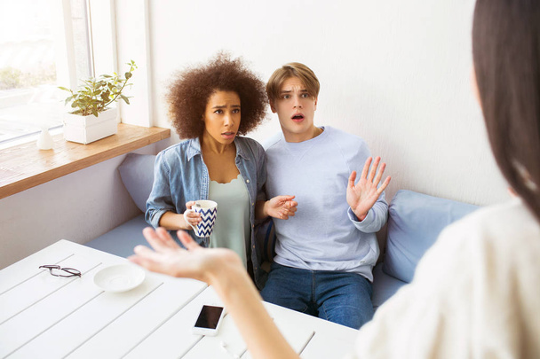 Girl in white sweater is standing in front of her friends and waving with her hand. Afro american girl and a guy are itting on sofa. They look confused sand unhappy. - 写真・画像