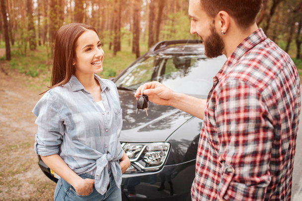 Nice and lovely couple is stansing near black car in forest. Guy and girl are looking to each other and smiling. He is giving her keys from car. - Fotografie, Obrázek