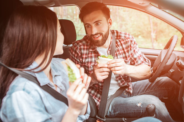 Nice and good-looking couple is sitting together in car and looking to each other. They are locked with the seatbelt. People are holding sandwiches. They made a stop to eat food. - Photo, image