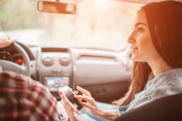 Girl is sitting with her boyfriend in car and looking at him. They are travelling. She is holding a phone is her hands and looking at guy. - Foto, Imagem
