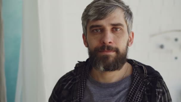 Close-up portrait of handsome bearded man with brown eyes and grayish hair wearing casual clothes and looking at camera. People and interior concept. - Imágenes, Vídeo
