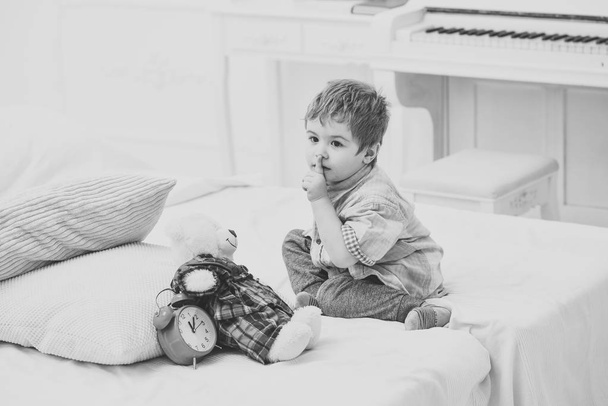 Child in bedroom with silence gesture. Kid put plush bear near pillows and alarm clock, luxury interior background. Time to sleep concept. Boy with calm face puts favourite toy on bed, time to sleep - Fotoğraf, Görsel