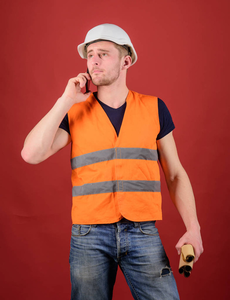 Architect calling engineer to fix plan. Communication concept. Man, foreman in helmet speaking on phone, red background. Engineer, architect, on busy face speaks on smartphone while holds blueprints - Photo, image